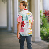 This outdoor photo shows a young man from the back and side, wearing a short-sleeved shirt with a bold tropical floral pattern in shades of pink, red, purple, and teal on a white background. He is standing under a covered walkway lined with white columns, looking to his left, and wearing dark pants.