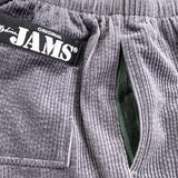 This is a close-up image of gray corduroy shorts, focusing on the texture and details. A black patch with "ORIGINAL JAMS" in white lettering is visible on the upper left, and a pocket opening reveals a dark green lining.
