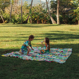 Two children, a boy and a girl, are sitting and interacting on a bright, patterned picnic blanket on a large, manicured lawn surrounded by tropical trees and distant mountains under a clear sky.