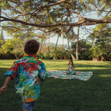 A boy, wearing a colorful shirt matching the blanket, is walking toward the camera with his back to the viewer, while a girl sits quietly on the patterned blanket far in the background beneath the wide, spreading branches of a large tree.