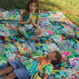 Two children are lying or sitting on a vibrant patchwork blanket spread on the grass; the girl is sitting cross-legged and drinking from a juice box, while the boy is lying on his back, and both are wearing matching tops with the blanket's colorful pattern.