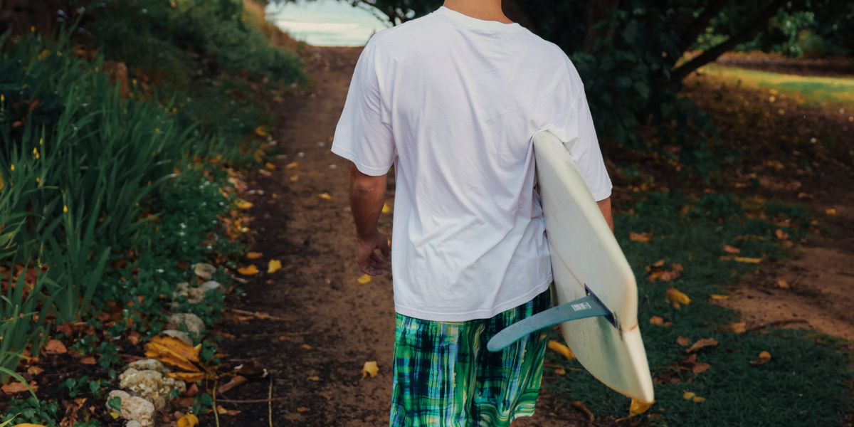 T-shirt blanc, short à carreaux verts et bob assorti, planche de surf à la main, marchant vers la plage.
