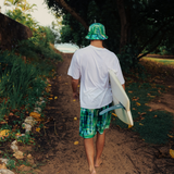 White tshirt with green plaid shorts and matching bucket hat holding surfboard walking towards the beach.