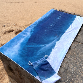 beach towel with picture of surfer surfing a big wave.  Towel is laying on the beach rocks and sand 