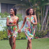 Two women walking hand-in-hand outdoors on a grassy lawn, wearing matching colorful floral slip dresses in a tropical garden setting.
