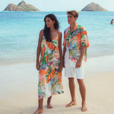 Dos personas con coloridos atuendos playeros de pie en una playa de arena con el océano y las islas al fondo.