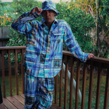 Person wearing a blue plaid shirt and pants on a wooden deck with greenery in the background