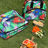 Colorful floral-patterned cooler bag on grass with snacks and fruits.