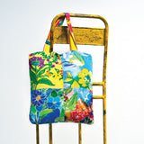 Un bolso de tela con asas estampadas multicolores cuelga de una silla o escalera de metal amarillo brillante y desgastado. El bolso presenta un mosaico de cuatro cuadrados con estampado floral vibrante en colores como amarillo, azul, rojo, morado y verde. El fondo es una pared blanca.