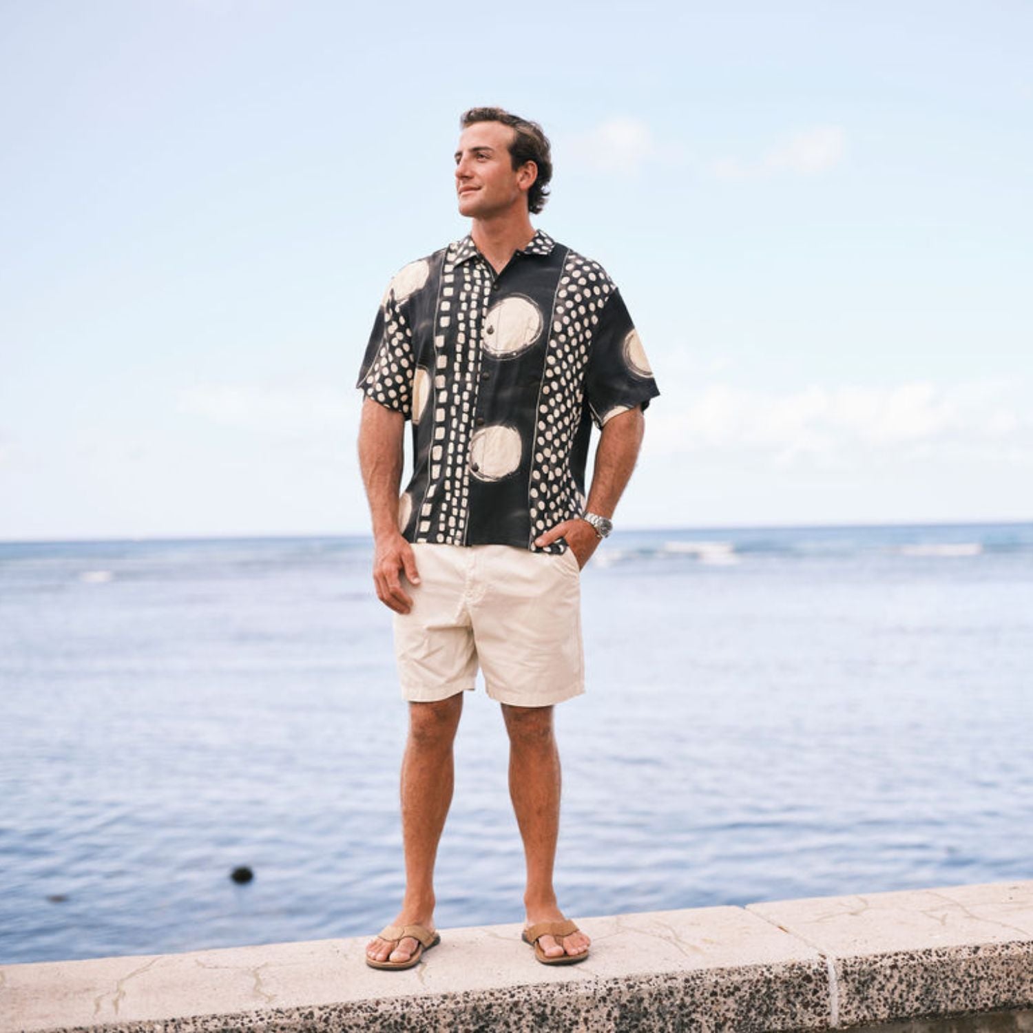 Man wearing a patterned shirt and shorts standing by the water.