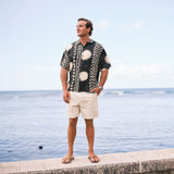 Man wearing a patterned shirt and shorts standing by the water.