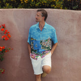 Un homme portant une chemise colorée à motif paysager se détache sur un mur rose orné de fleurs rouges.