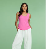 A smiling woman with long dark hair is posing confidently against a pale mint green background. She is wearing a bright pink scoop-neck sleeveless tank top tucked into high-waisted, flowy white wide-leg pants. Her hands are placed casually in the pockets of the pants.