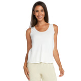 Una donna sorridente con lunghi capelli scuri è in piedi su uno sfondo bianco, indossando una canotta bianca senza maniche con orlo leggermente svasato e pantaloni beige chiaro e fluidi. La canotta ha una semplice scollatura a barchetta.