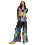 A woman with long dark hair stands facing the camera, wearing a matching resort set against a white background. The set consists of a black short-sleeve collared shirt and flowing wide-leg pants. Both pieces feature a large, colorful tropical print of overlapping leaves in neon pink, turquoise, lime green, and burnt orange. She is wearing woven straw platform sandals.