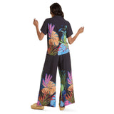 The back view of a woman wearing the black short-sleeve shirt and wide-leg pants set with the colorful tropical print. The large print is prominently displayed across the back of the shirt and on the lower legs of the pants. She is standing with her arms slightly raised, and she is wearing woven straw platform sandals.