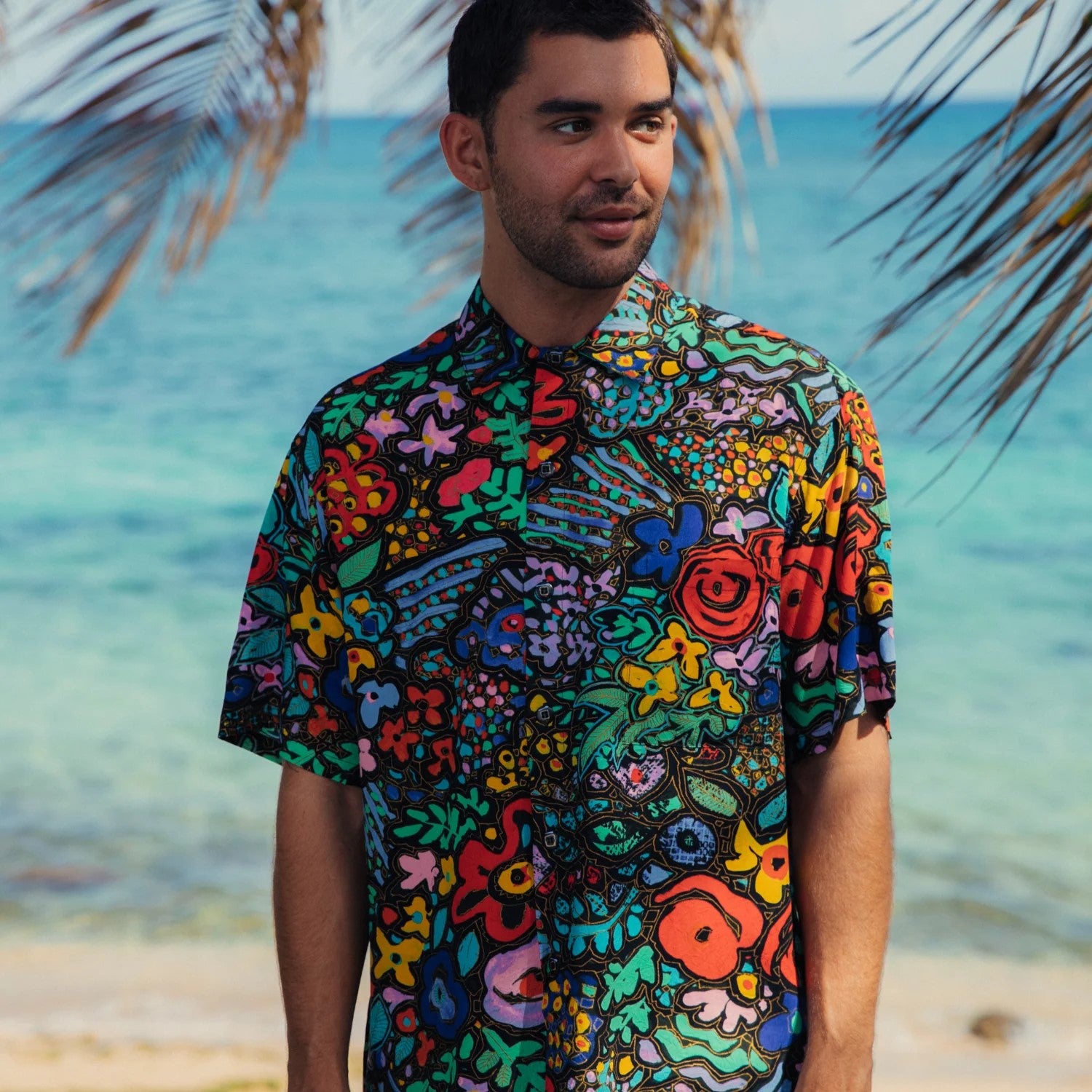 Man wearing a colorful patterned shirt standing on a beach with palm leaves and ocean in the background