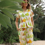 Femme vêtue d'une tenue colorée à motifs de poires, debout à l'extérieur, avec de la verdure en arrière-plan.