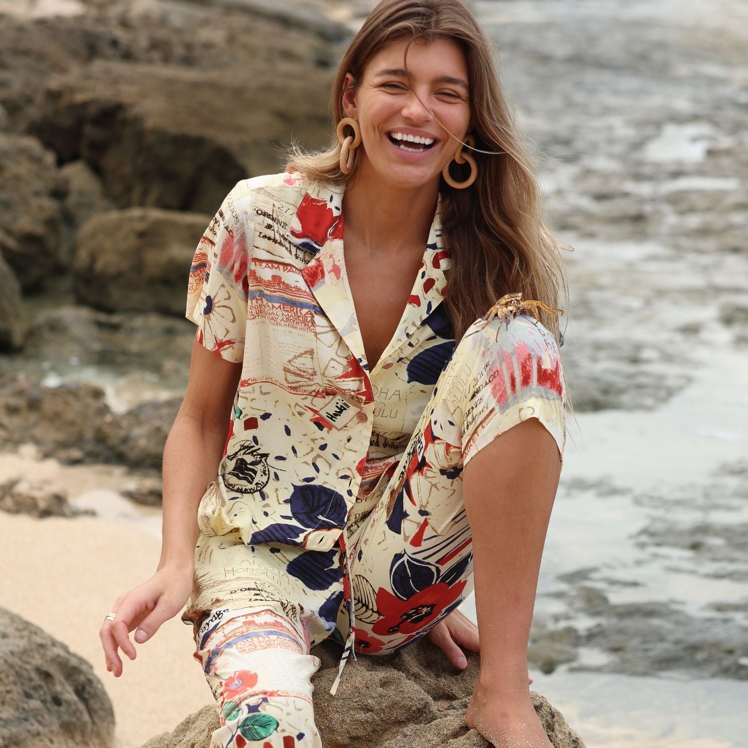 A smiling woman with long blonde hair is sitting barefoot on a large rock on a beach. She is wearing a matching short-sleeve collared shirt and cropped pants set, featuring a vintage travel-themed print in cream, red, and navy blue. She is wearing large, circular wooden earrings.