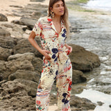 A woman in a matching vintage travel-print shirt and cropped pants is standing on a rocky beach, looking toward the side. Her hands are resting in the pockets of the pants, and the background shows a shallow tide pool and large grey boulders.
