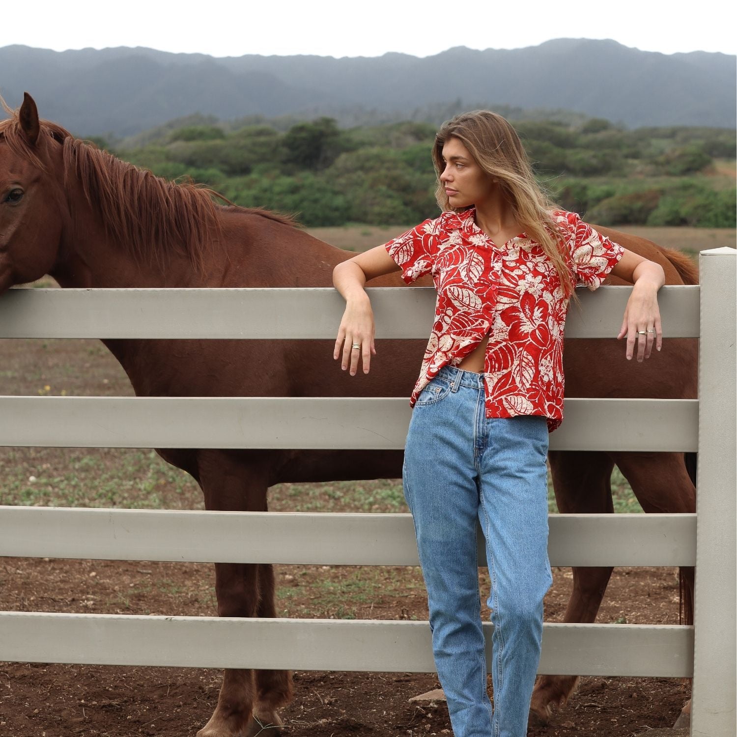 Une femme, vêtue d'une chemise à imprimé tropical rouge et blanc sur un jean clair, est debout, appuyée contre une clôture en bois blanc. Un cheval brun se tient juste derrière elle, à gauche du cadre. À l'arrière-plan, on aperçoit des collines verdoyantes et des montagnes lointaines sous un ciel nuageux.