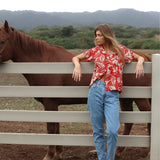 Une femme, vêtue d'une chemise à imprimé tropical rouge et blanc sur un jean clair, est debout, appuyée contre une clôture en bois blanc. Un cheval brun se tient juste derrière elle, à gauche du cadre. À l'arrière-plan, on aperçoit des collines verdoyantes et des montagnes lointaines sous un ciel nuageux.