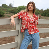 Person wearing a red floral shirt and blue jeans leaning against a fence with a rural background.