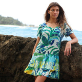 Woman in a colorful dress with leaf pattern leaning against rocks by a beach.