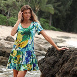 A woman wearing a short-sleeve mini dress with a tiered ruffle skirt is posing on a rocky shoreline. The dress features an abstract tropical leaf print in shades of bright blue, forest green, and yellow. She is leaning on a dark rock with one hand and is looking off to the side. The background shows a sandy beach and palm trees.