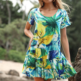 A mid-chest view of a woman on the beach, highlighting the short-sleeve mini dress and its print. The dress has a scoop neckline and a tiered ruffle skirt, all covered in a colorful abstract print of blue, green, and yellow tropical leaves. She is holding her hand on her hip and the beach is visible behind her.