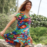 A woman with long blonde hair is smiling and appears to be twirling on a rocky beach, causing the ruffled skirt of her dress to lift slightly. She is wearing bright red earrings and a short-sleeve dress with a multicolored, abstract fish print. The setting is sunny with green foliage and palm trees in the background.