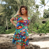 A woman with long blonde hair and bright red earrings is standing on a rocky beach, looking off to the side. She is wearing a short-sleeve mini dress with a tiered ruffle skirt, featuring an extremely colorful, abstract print of fish and underwater brushstrokes in neon blue, green, red, yellow, and black. Palm trees and foliage are visible in the background.