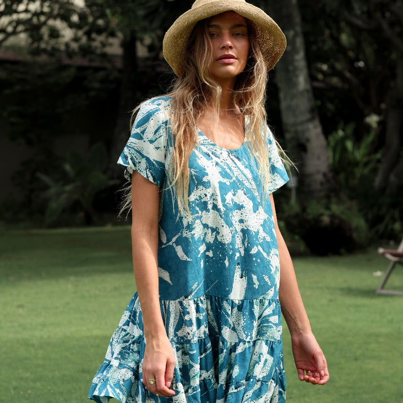 A woman with long, sun-kissed hair is standing outdoors on a green lawn, looking directly at the camera. She is wearing a woven straw hat and a short-sleeve mini dress with a scoop neck and tiered ruffle skirt. The dress has an abstract, batik-style print in deep teal and white. Palm trees are visible in the blurred background.