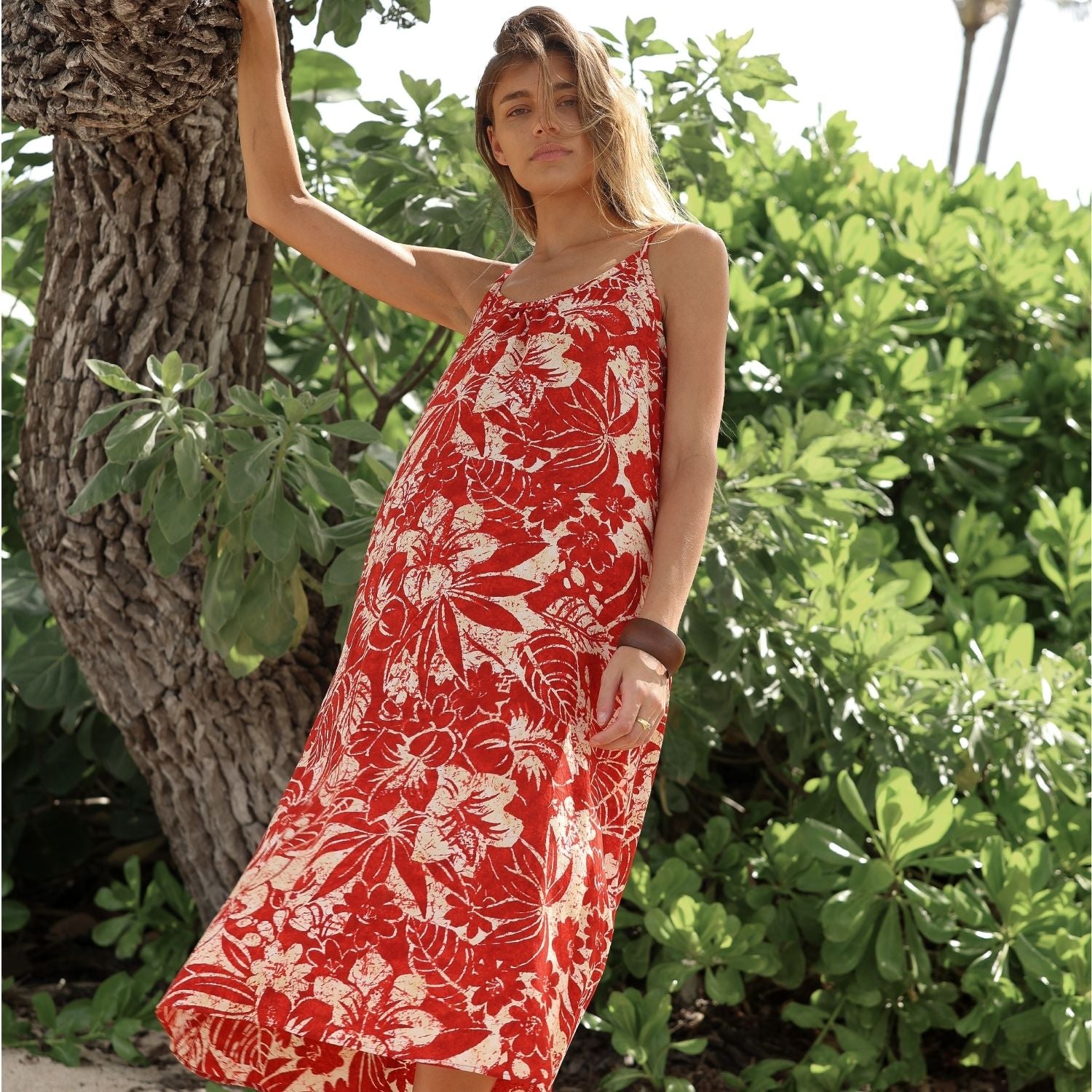 Femme vêtue d'une robe rouge à fleurs, debout au milieu d'un feuillage vert.