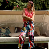 Woman in a pink top and colorful pants sitting on a bench with greenery in the background