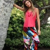 A woman stands against a large tree trunk wearing a vibrant pink tank top and wide-leg trousers with a bold, colorful abstract print.