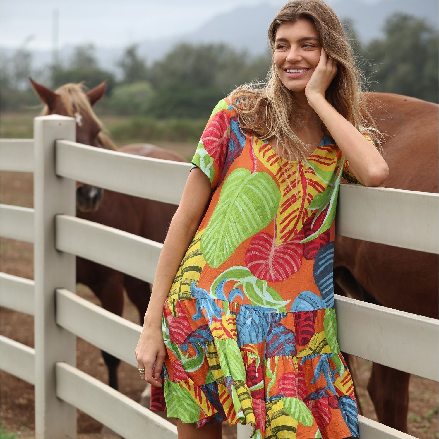 In a candid-style shot, the same woman from the previous image is seen smiling and looking off-camera. She is leaning her elbow on the top rail of a white fence, resting her face in her hand. The colorful tropical dress has a tiered, ruffled skirt. Behind her, a brown horse is partially visible, and the landscape behind them suggests a wide, open ranch or farm.