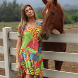 A young woman with long light-brown hair stands outdoors in a rural setting. She is wearing a V-neck, knee-length dress featuring the colorful tropical leaf pattern from the previous image. She leans her arm on a white, three-rail PVC fence, with a reddish-brown horse standing directly behind her, its head resting near her shoulder. The background shows a soft-focus field and trees under an overcast sky.