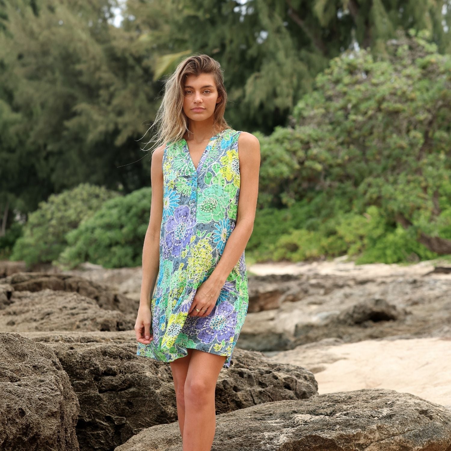 A woman standing among large beach rocks wearing a colorful floral batik dress, with a forest and sandy beach in the background.