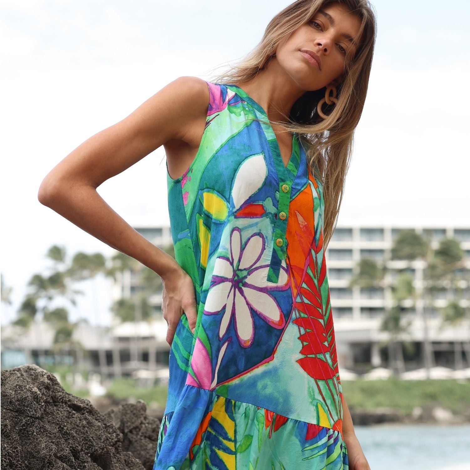 A woman posing in a colorful tropical sleeveless dress against a coastal resort background.