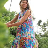 A woman smiling and looking away while wearing a colorful bohemian-print sleeveless dress in a tropical outdoor setting.
