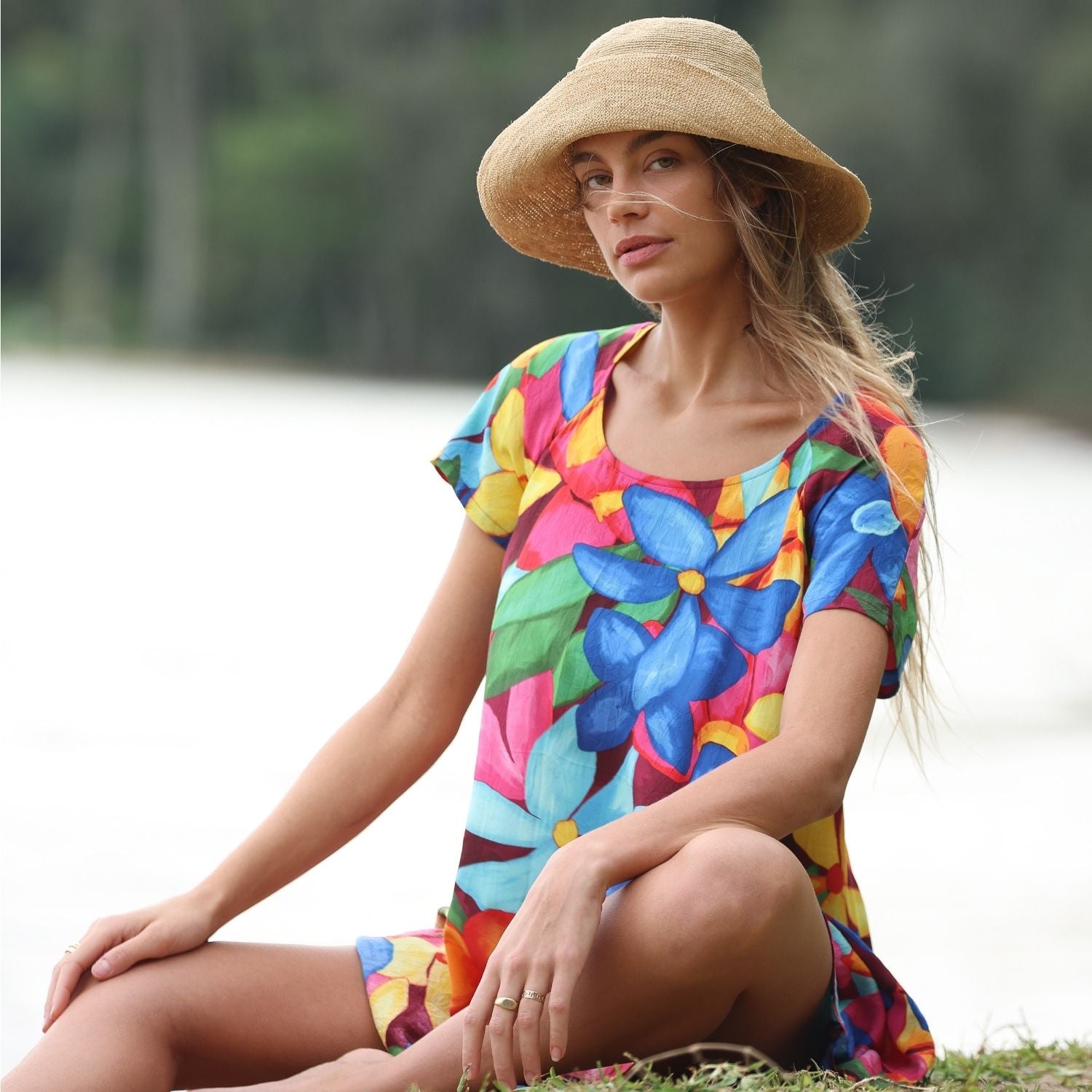 Woman wearing a colorful floral dress and straw hat sitting outdoors.