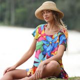 Woman wearing a colorful floral dress and straw hat sitting outdoors.