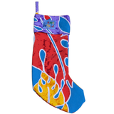 A christmas stocking featuring a bold, abstract print in primary colors. The main body of the stocking is covered in large shapes of red, blue, and yellow with white outlines details, resembling splatters or leaves. The cuff is a contrasting purple fabric with a subtle blue pattern and a small blue patch displaying the "Jams World" logo.