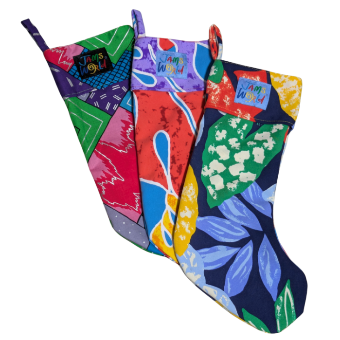 Three brightly colored christmas stockings are grouped together. The left stocking features a patchwork geometric pattern in shades of purple, green, and pink. The middle stocking has a red, blue, and yellow abstract print. The right stocking features large blue and green tropical leaves on a dark blue background. All three have a small "Jams World" logo on the cuff.