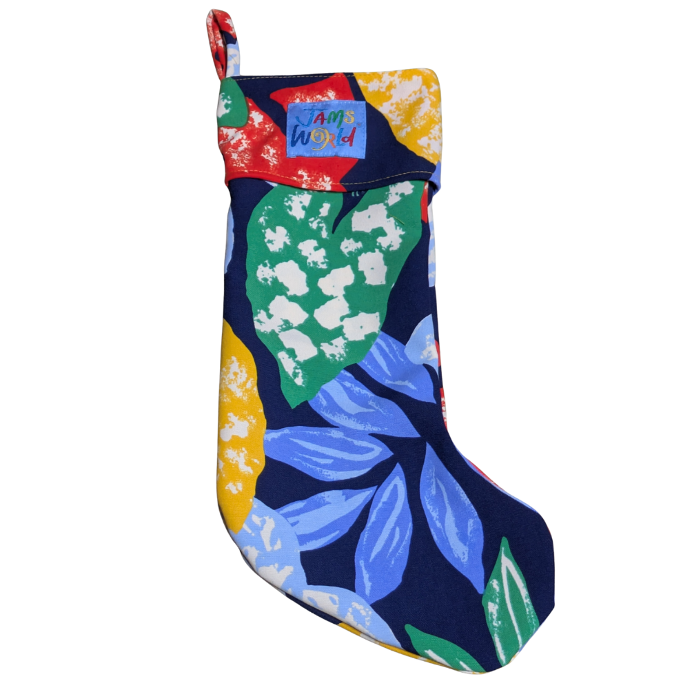 A single christmas stocking featuring a vibrant, abstract tropical leaf pattern in bright colors. The dark blue fabric is covered with large leaves in shades of blue, green, yellow, and white. The stocking has a wide cuff made from the same print, with a small blue "Jams World" logo patch sewn onto it.