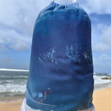 A close-up of a small, cylindrical drawstring pouch featuring a blue ocean and white foam print, with a beach and cloudy sky in the background.
