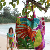Un bolso de tela teñido con la técnica tie-dye, de colores vivos, cuelga de la rama de un árbol, con una persona borrosa vestida con un vestido colorido al fondo.