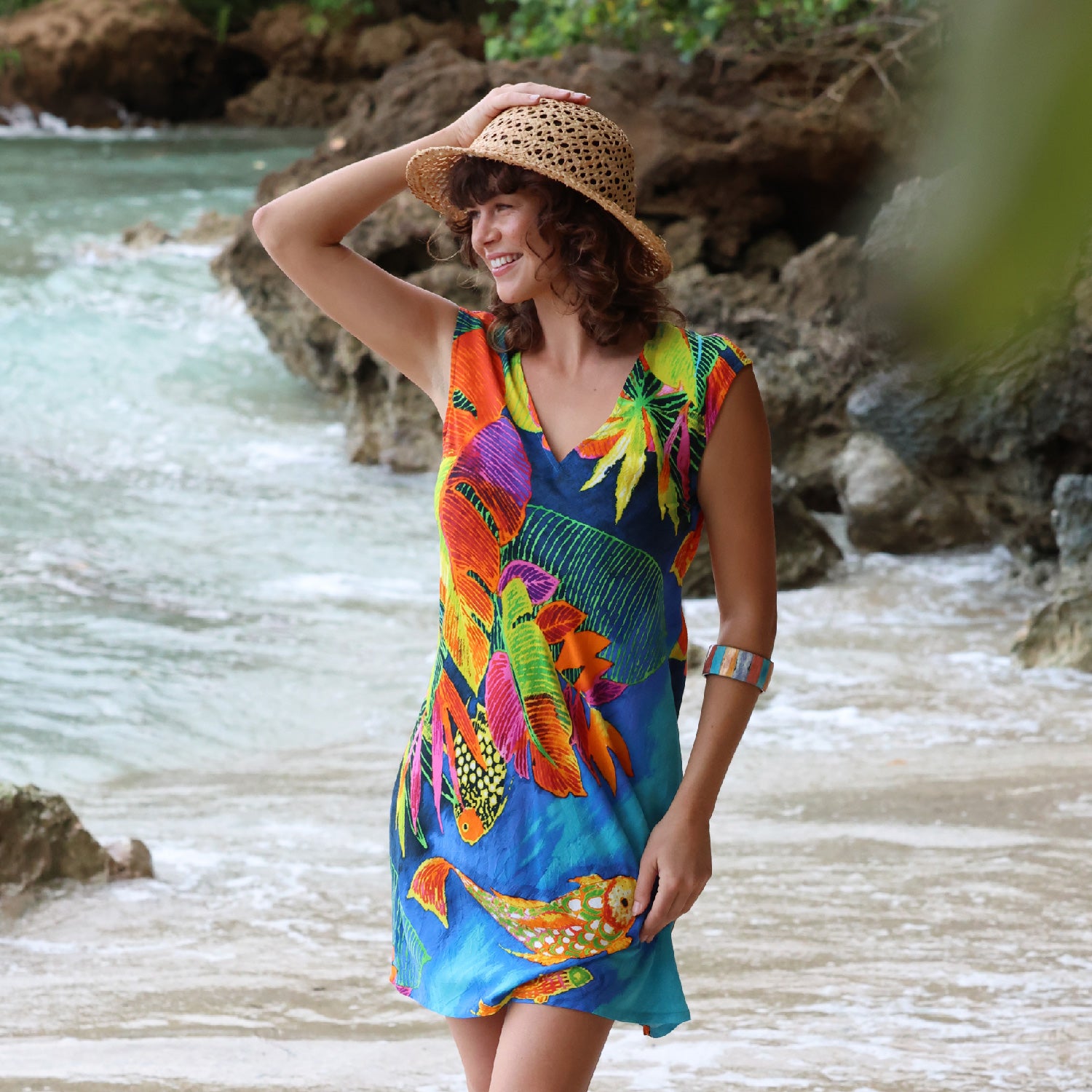 Une femme en robe colorée et chapeau de paille se tient au bord d'une rivière avec des rochers et de l'eau.