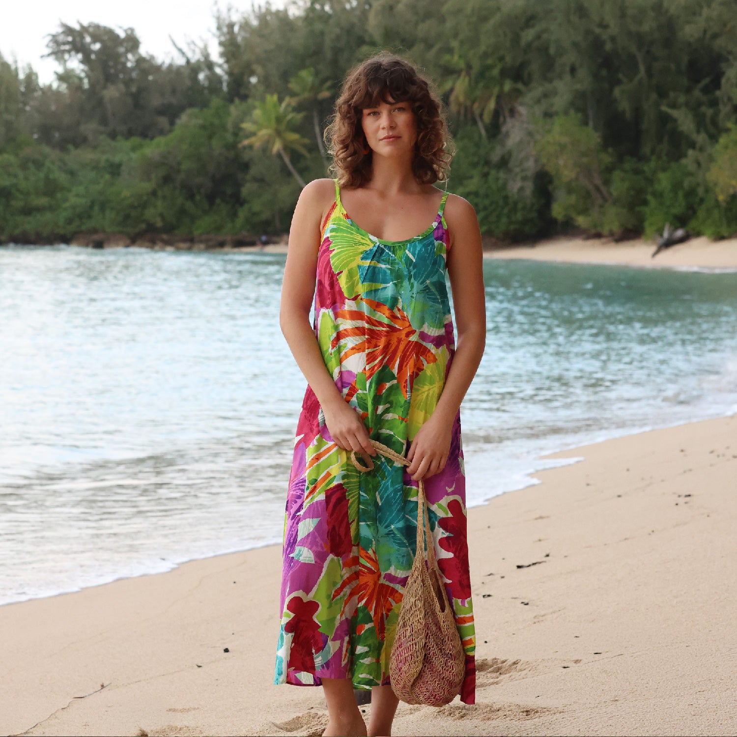 Woman in a colorful dress standing on a beach with ocean and trees in the background